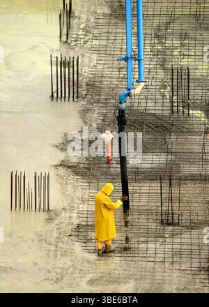 Construction worker in yellow waterproof poncho pouring concrete from a pump at a building site, standing with feet in fresh wet concrete Stock Photo