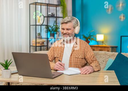 Caucasian mature man sitting at home table listening to podcast or online stream on laptop with headphones. Guy takes making notes into notebook, interested engaged in content, learning education Stock Photo