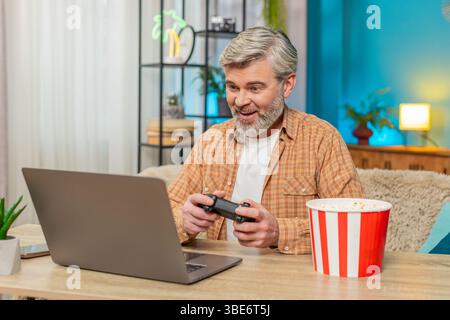 Caucasian mature man playing video games on laptop with gamepad joystick. Middle-aged guy sitting at home table eats popcorn, looks engrossed enjoys gaming experience showing dedication and enthusiasm Stock Photo