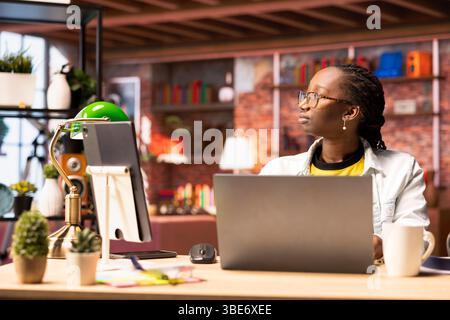 Contemplative woman daydreaming, gazing outside the window, feeling bored after working too much on job tasks. African american person at home caught in thoughts, pondering Stock Photo