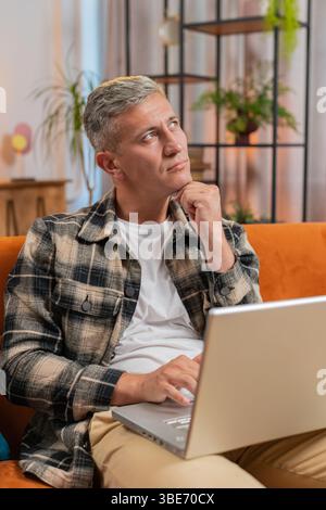 Caucasian man freelancer with gray hair working on laptop computer with pensive expression, pondering a solution answer, doubting question, sends messages, makes online shopping purchases at home room Stock Photo