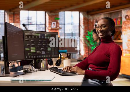 Portrait of upbeat woman working in innovative startup office using programming languages to build application. Jolly African american software development agency IT expert optimizing code on computer Stock Photo