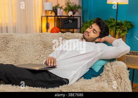 Indian young man at home, exhausted from work, falling asleep on the sofa with a laptop on his lap. Arabian guy tired lazy after long hours of work, resting on the couch, overwhelmed and drained Stock Photo