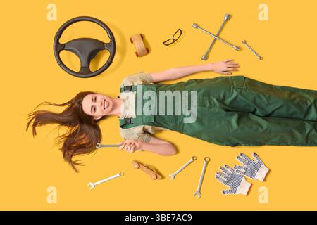 Happy young female auto mechanic with different tools and steering wheel lying on yellow background Stock Photo