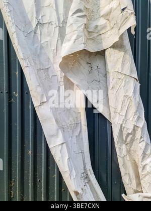 Close up of a weathered and torn white tarp hanging against a dark green corrugated metal wall, showing signs of age and exposure to the elements Stock Photo