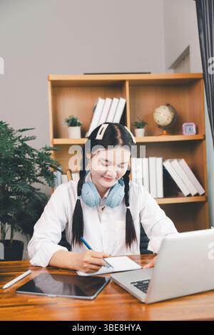 Asian girl student wear headphones learning online watching webinar class looking at laptop computer elearning remote lesson making notes or video cal Stock Photo