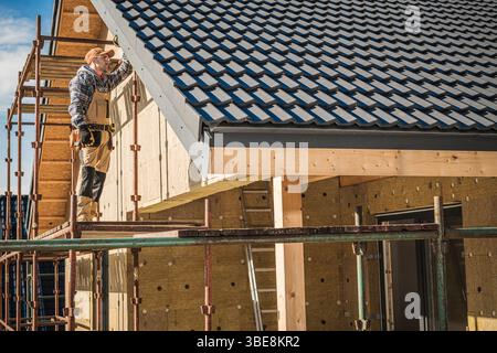 A construction worker is carefully placing roof tiles on a house being built. Scaffolding surrounds the building, with tools at hand and a bright sky Stock Photo