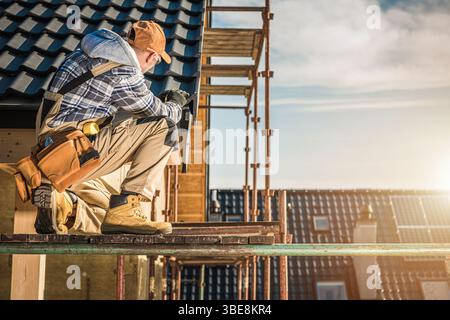 A construction worker kneels on scaffolding, checking tools and materials at a building site as the sun sets, illuminating the rooftops around him. Stock Photo