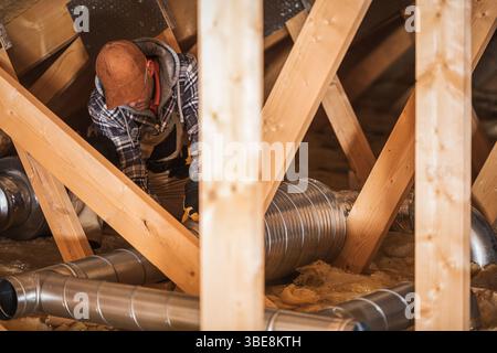 A worker is focused on repairing a ventilation system inside a residential attic, surrounded by wooden beams and insulation during daylight. Stock Photo