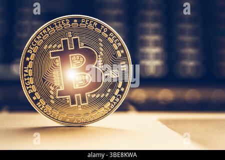 A Bitcoin coin stands upright on a table, showcasing its intricate design and gold color, while a blurred screen with cryptocurrency data offers conte Stock Photo