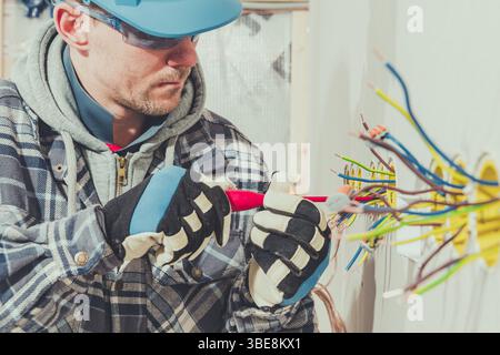 A technician focuses on connecting wires at a construction site, wearing protective gloves and safety glasses while working on electrical installation Stock Photo