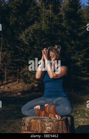 A woman practicing yoga performs a sitting in lotus position on a tree stump, meditating alone in casual clothes on a sunny autumn day in the park. Hi Stock Photo