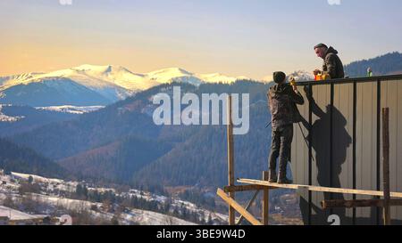 Construction workers building a house in the mountains. Two construction workers assembling a metal roof on a house, with a stunning backdrop of snow Stock Photo