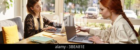 Two young women enjoy their time working remotely together in a stylish cafe setting, banner Stock Photo