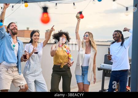 Friends dancing and drinking at rooftop party under string lights Stock Photo