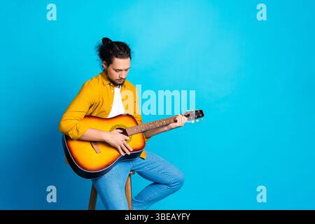 Young man plays acoustic guitar against a vibrant blue background showcasing casual style and musical passion Stock Photo