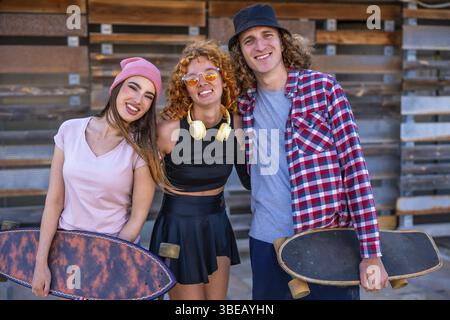 Three young skaters smiling and posing together with their skateboards in front of a rustic wooden wall, showcasing their friendship and style Stock Photo