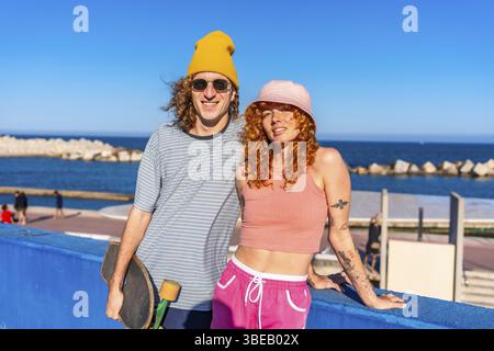 Young couple of skaters posing with their skateboard by the sea, enjoying a sunny summer day filled with love and laughter Stock Photo