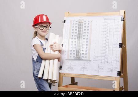 Girl Builder, architect, holding a bunch of drawings, standing at the blackboard with the drawing of the building Stock Photo