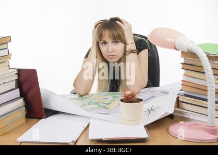 Student designer behind a desk with a pile of books and projects sits holding his head in his hands Stock Photo