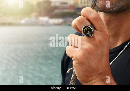 Bearded sailor displaying an elegant ring adorned with a golden anchor and rope design while gripping a rope in a bustling port, embodying the essence of maritime jewelry Stock Photo
