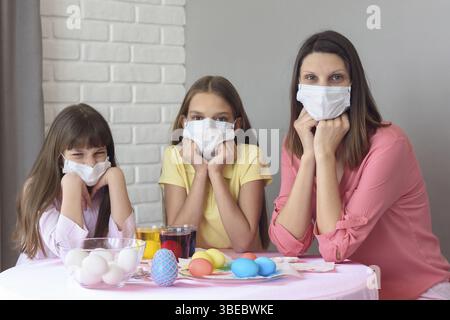 Mom and two daughters at the table in medical masks paint Easter eggs for the holiday Stock Photo