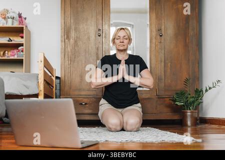 Middle age woman mediating and doing yoga at home with laptop online class. Stock Photo
