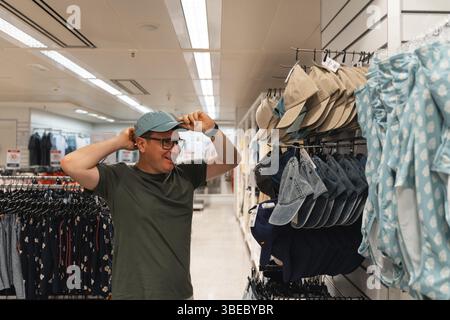 A man tries on a blue hat while browsing a selection of hats in a clothing store. Male consumerism, second hand shopping . Stock Photo