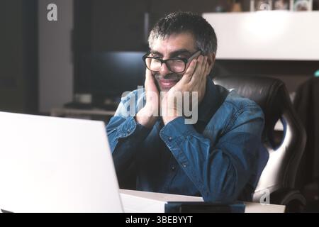 A smiling man sitting on a desk and looking at the laptop in front of him. He wears glasses and has his hands on his head. Business concept at home. Stock Photo