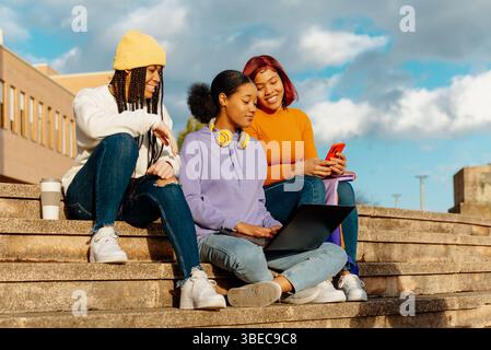 Three female students sitting together on campus stairs, engaged in study and conversation under a clear sky Stock Photo