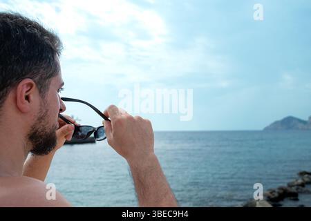 Young adult man taking off his sunglasses contemplating beautiful beach horizon and sunny blue sky Stock Photo