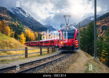 Red modern electric train traveling along a railway, winding through a stunning high mountains with snow-capped peaks and vibrant autumn trees in swit Stock Photo