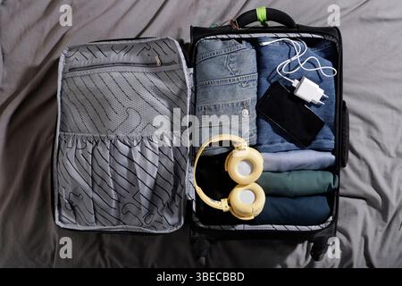carry-on suitcase with blue clothes and large headphones, charger and cell phone on the bed Stock Photo