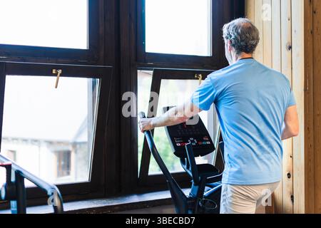 A man is exercising on a stationary bike in front of a window The man is wearing a blue shirt and white pants Stock Photo