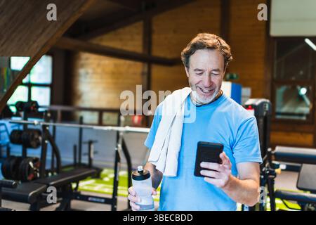 A man is smiling while looking at his cell phone in a gym He is holding a water bottle and a towel Stock Photo