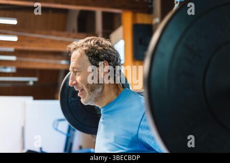 Portrait of a mature man lifts a barbell with weighted discs in a gym He is training his leg strength by performing squats He smiles and enjoys himsel Stock Photo