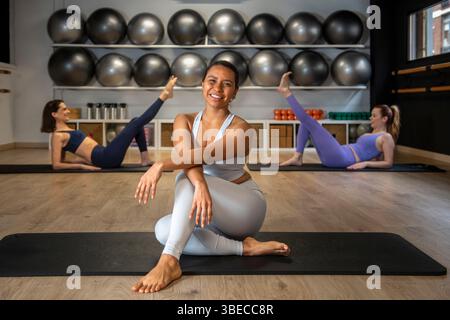Pilates instructor smiling and sitting on mat while her students are exercising on the floor in a gym Stock Photo