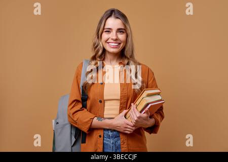 A cheerful student with long hair beams joyfully while holding a stack of books. Dressed casually yet stylishly, she carries a trendy backpack, ready Stock Photo