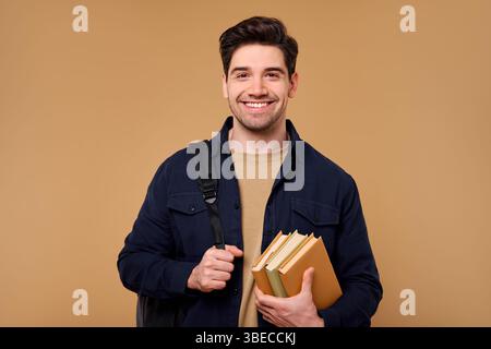 A joyful young man, holding a tall stack of colorful books and wearing a stylish backpack, stands confidently against a beige background, exuding posi Stock Photo