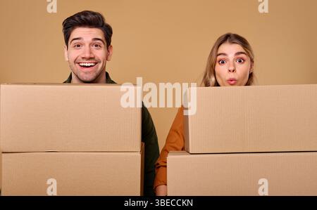 A cheerful couple is smiling broadly as they pose playfully behind a stack of cardboard boxes, which clearly conveys their excitement about moving or Stock Photo