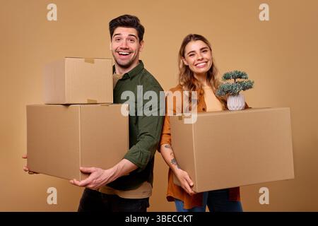 A cheerful young couple is depicted standing backtoback, each holding cardboard boxes as they prepare for an exciting move. The warm and vibrant backg Stock Photo