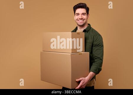 A cheerful man confidently standing, carrying two cardboard boxes. The warm beige background enhances the positive vibe and highlights his activity of Stock Photo