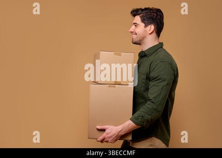 A cheerful man confidently standing, carrying two cardboard boxes. The warm beige background enhances the positive vibe and highlights his activity of Stock Photo