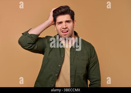 A young man, puzzled and frustrated with his hand on his head against a solid background, conveys deep confusion about a challenging dilemma, leaving Stock Photo