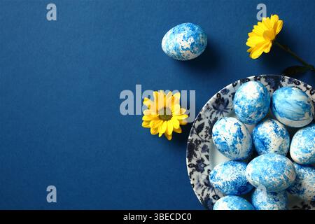 Plate with blue and white Easter eggs and yellow flowers on dark blue table. Top view. Stock Photo