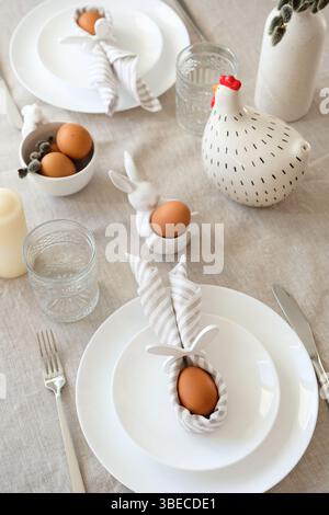 Easter dining table setting with Easter bunny made of linen napkin and egg on plate, cutlery. Stock Photo