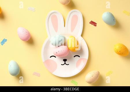 Happy Easter card. Bunny shaped plate with colorful Easter eggs on pastel yellow background. Flat lay. Top view. Stock Photo