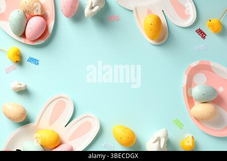 Easter frame border made of colorful Easter eggs, bunny and eggs shaped cutlery on pastel blue table. Stock Photo