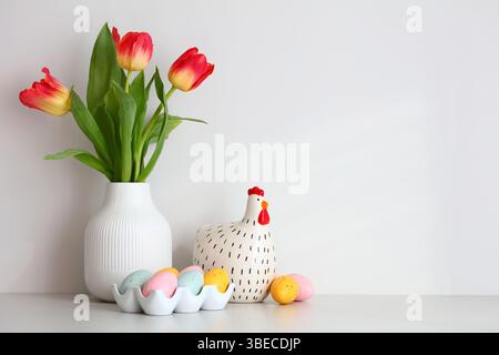 Vase of tulips, decorative chicken, colorful Easter eggs on table in scandinavian room interior. Stock Photo