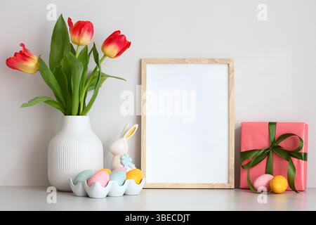 Empty vertical picture frame mockup with vase of tulips, colorful Easter eggs, gift box on table in scandinavian room interior. Stock Photo